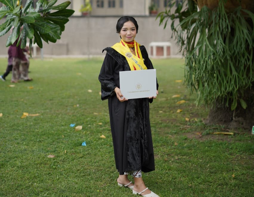 Graduate in a black gown with a yellow stole, holding a diploma outdoors on campus.