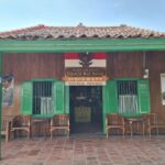 Front view of a rustic wooden Indonesian house with green trim, open doorway, and wicker chairs on a tiled veranda beneath a red-tiled roof.