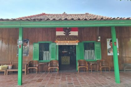 Front view of a rustic wooden Indonesian house with green trim, open doorway, and wicker chairs on a tiled veranda beneath a red-tiled roof.