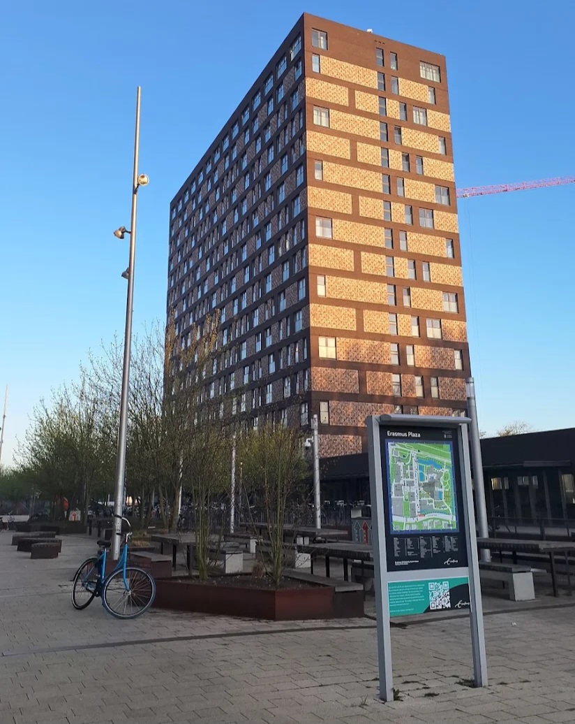 Tall brick apartment building with many windows in a city plaza, bike resting on a rack, and an Erasmus Plaza map sign in the foreground.