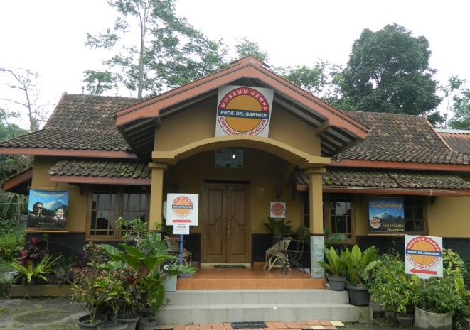 uopp - Mabur.co Front view of a small museum building with yellow walls, red-tiled roof, and a circular sign above the entrance.