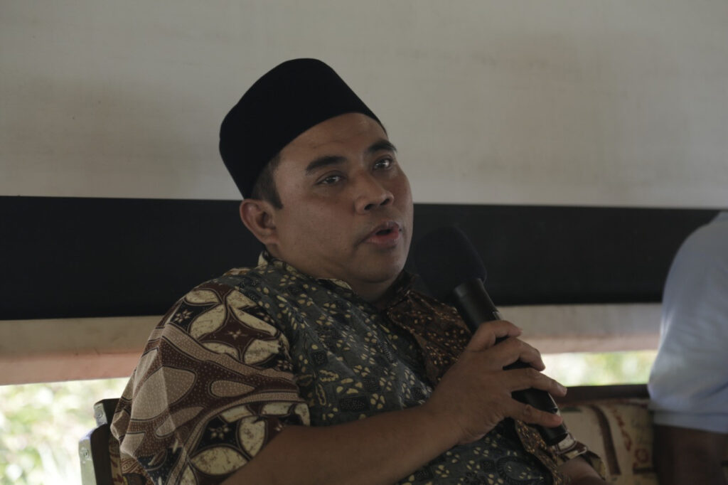 uscuacoxsa - Mabur.co Man wearing a batik shirt and black cap speaks into a handheld microphone while seated.