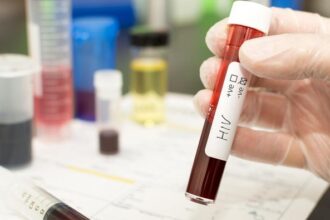 Gloved hand holding a blood sample tube labeled HIV in a laboratory, with other lab equipment blurred in the background.
