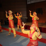 Dancers in red and yellow traditional costumes performing a synchronized routine on a gym stage.