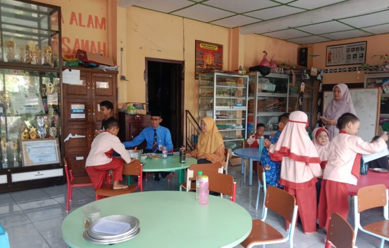 School cafeteria with round green tables, children in red-and-white uniforms eating; trophy cases along the wall to the left.