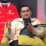 Man in a beige jacket speaks into a podcast microphone, seated on a yellow chair with a red soccer jersey and trophies behind him.