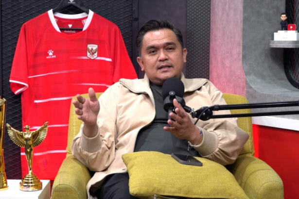 Man in a beige jacket speaks into a podcast microphone, seated on a yellow chair with a red soccer jersey and trophies behind him.
