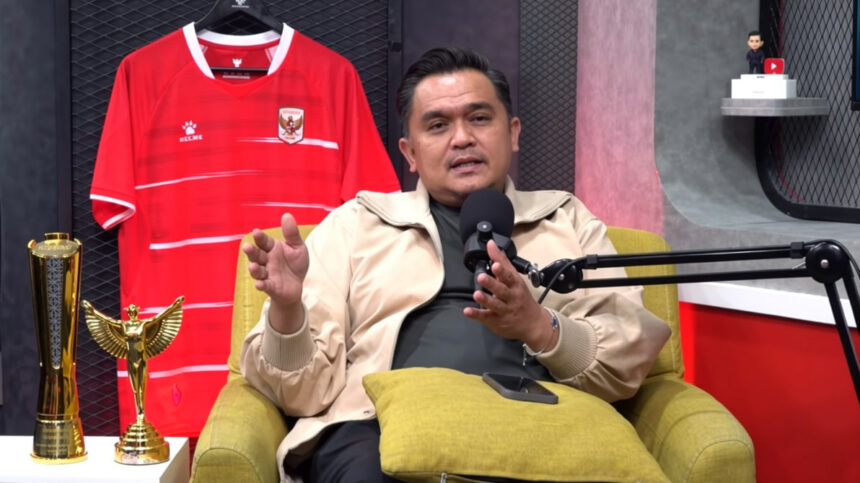 Man in a beige jacket speaks into a podcast microphone, seated on a yellow chair with a red soccer jersey and trophies behind him.