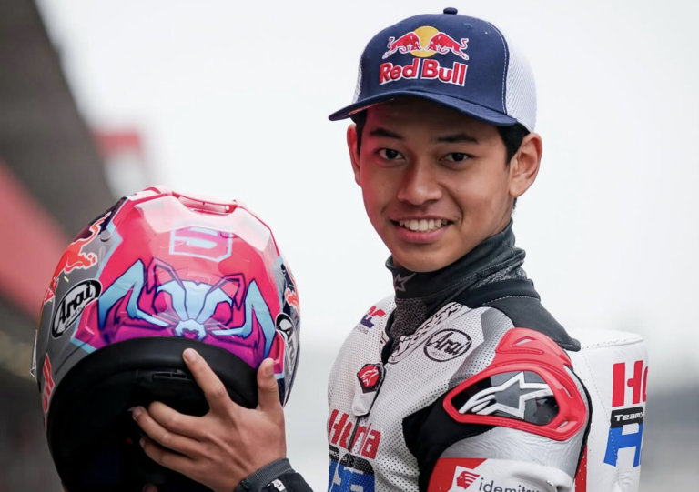 Motorsport rider in a white racing suit with red and black accents, smiling while holding a pink and blue helmet with sponsor logos, Red Bull cap in the background.
