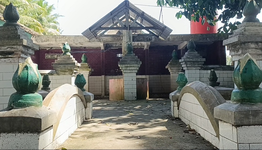 Stone bridge railing with green decorative finials leading to a small open pavilion in a sunlit temple courtyard.