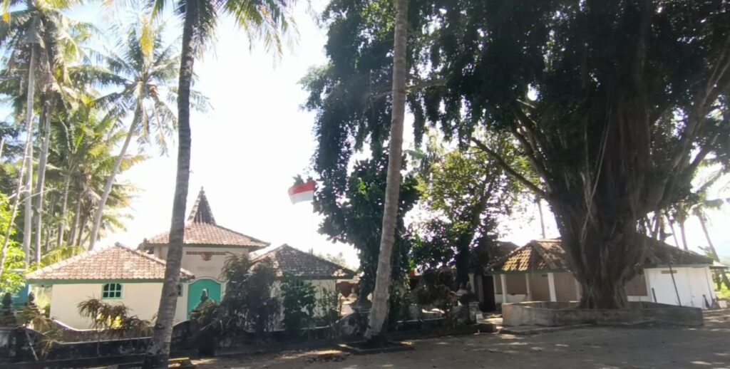 Tropical village scene with white houses, tiled roofs, and tall palm trees; an Indonesian flag is visible among the trees.