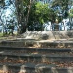Circular stone amphitheater-like steps leading up to a small monument statue among trees in a park setting