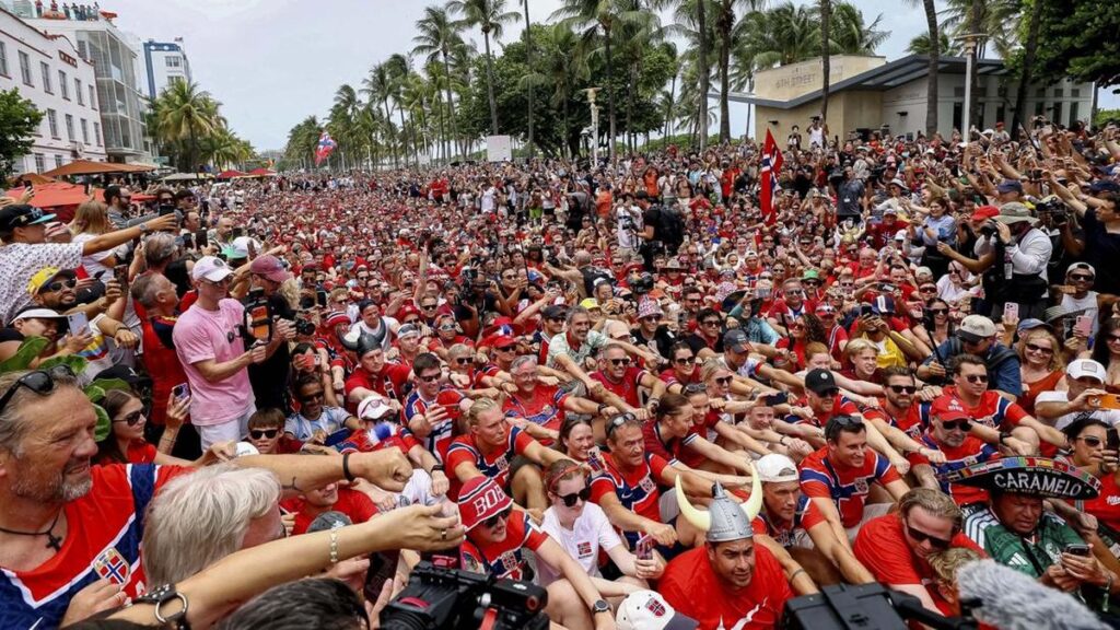 Huge crowd packed along a city street, mostly in red shirts, with palm trees and spectators on both sides, many people taking photos and raising arms.