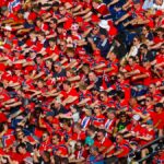 Large crowd of sports fans in red, seated in stadium stands and cheering at a game.