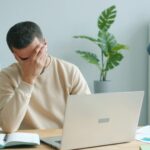 Man at a desk with a laptop, head in his hand, showing frustration in a bright home/office space with a plant in the background.