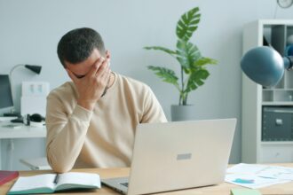 Man at a desk with a laptop, head in his hand, showing frustration in a bright home/office space with a plant in the background.