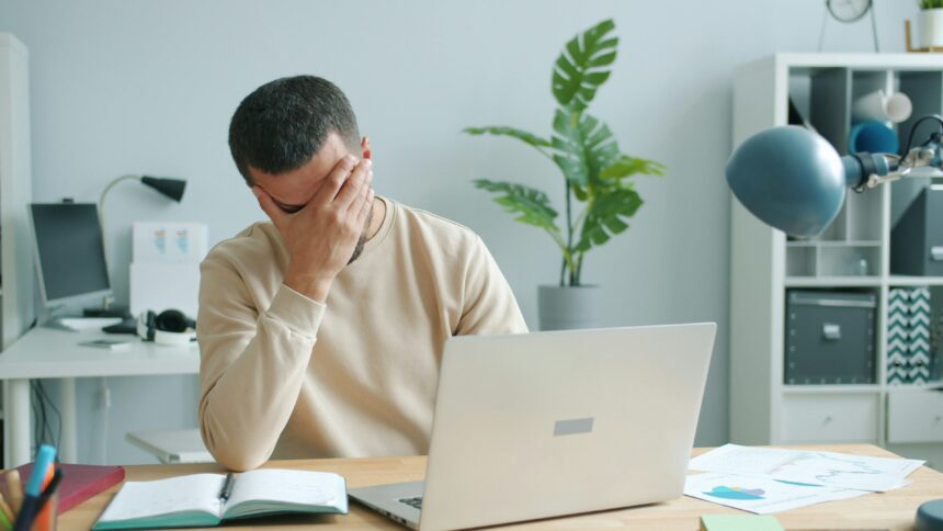 Man at a desk with a laptop, head in his hand, showing frustration in a bright home/office space with a plant in the background.