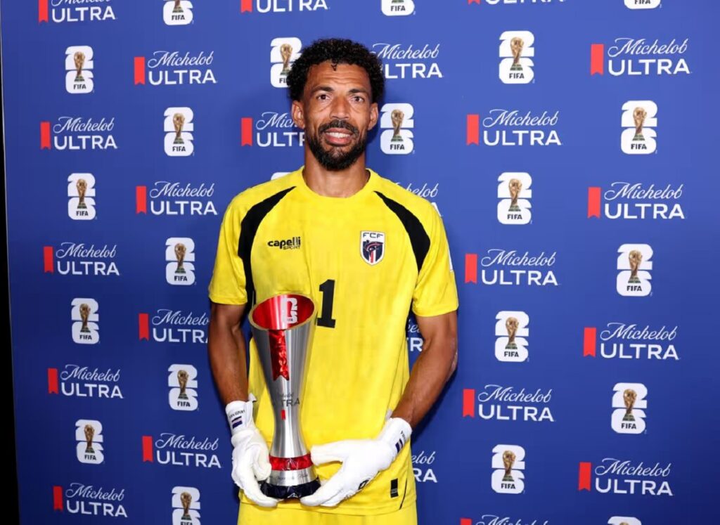 Soccer goalkeeper in a yellow kit holding a silver trophy, standing in front of a blue Michelob Ultra/FIFA backdrop.