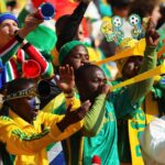 Energetic South African soccer fans in green and yellow cheer, blowing vuvuzelas.