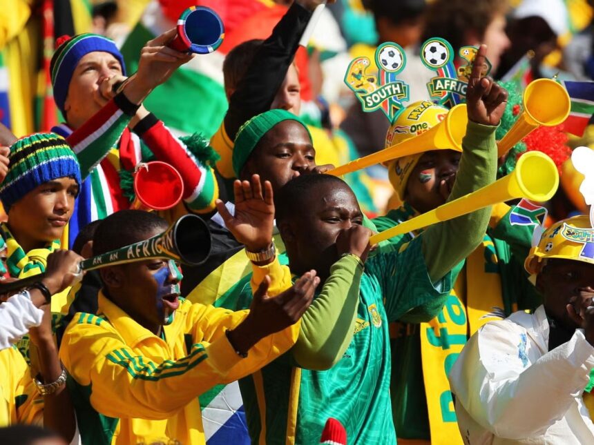 Energetic South African soccer fans in green and yellow cheer, blowing vuvuzelas.