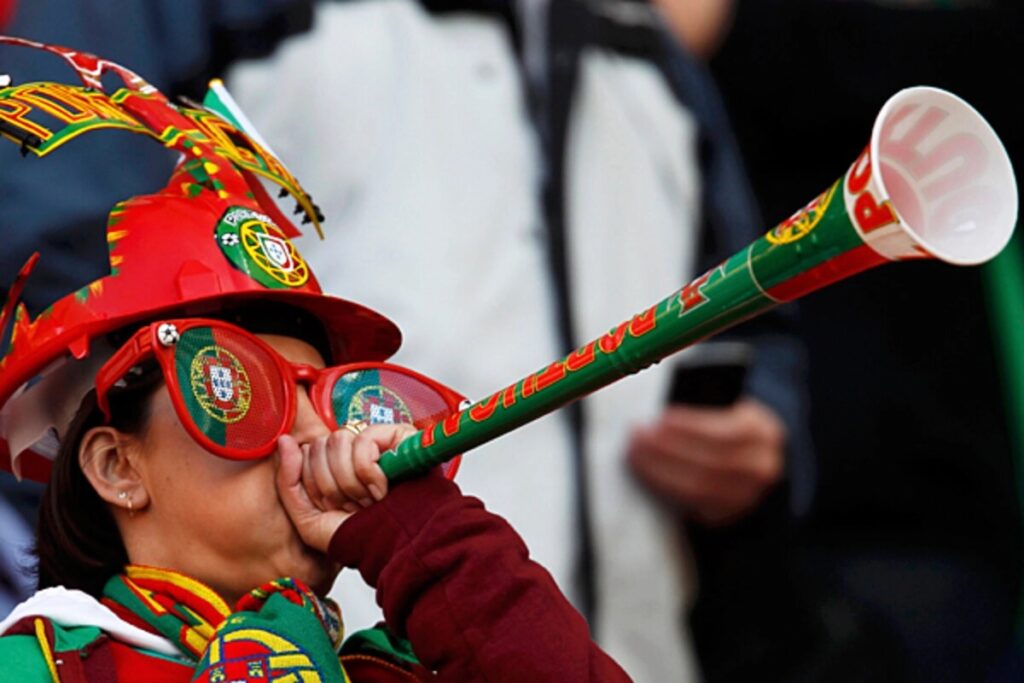 Fan dressed in Portugal colors blows a green and red novelty horn at a game, wearing red sunglasses and a festive hat.