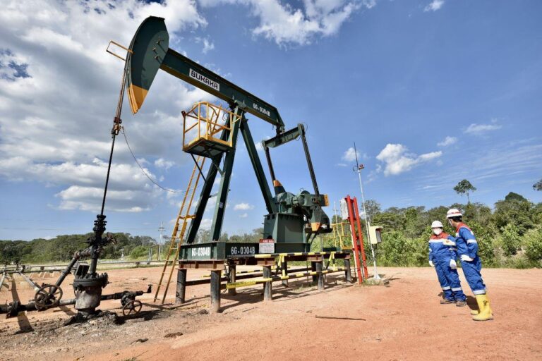 Oil pumpjack labeled Buraka in a dusty lot, with two workers in blue coveralls and hard hats nearby under a blue sky.