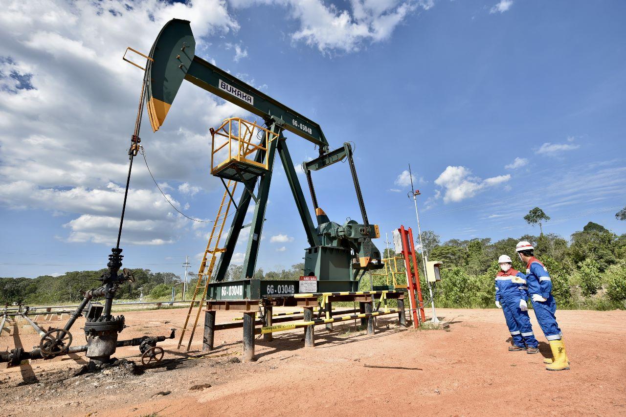 Oil pumpjack labeled Buraka in a dusty lot, with two workers in blue coveralls and hard hats nearby under a blue sky.
