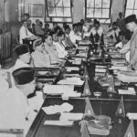 Crowded meeting around a long table with many attendees, a man stands at the right addressing the group amid papers and triangular numbered markers on the table.