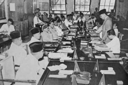 Crowded meeting around a long table with many attendees, a man stands at the right addressing the group amid papers and triangular numbered markers on the table.