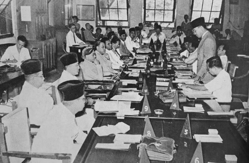Crowded meeting around a long table with many attendees, a man stands at the right addressing the group amid papers and triangular numbered markers on the table.