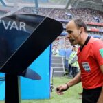 Football referee in a red shirt with a headset stands by a large video monitor on the sideline during a match.