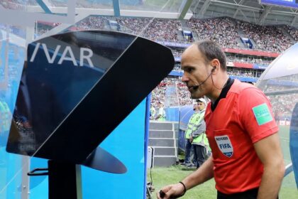 Football referee in a red shirt with a headset stands by a large video monitor on the sideline during a match.