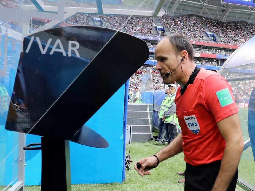 Football referee in a red shirt with a headset stands by a large video monitor on the sideline during a match.