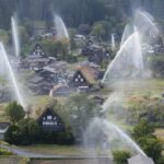 Aerial view of a rural village with steep thatched-roof houses as large water sprinklers arc over the scene, spraying fountains across the landscape.