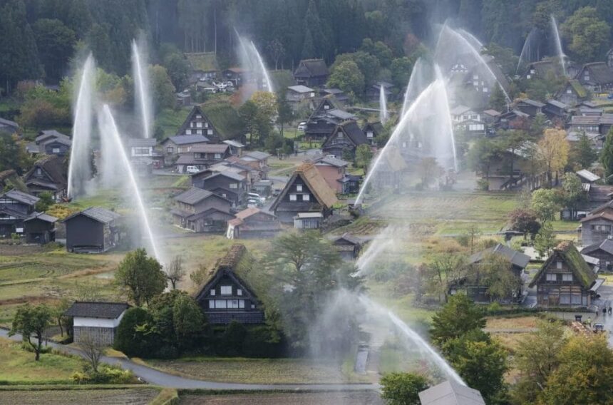 Aerial view of a rural village with steep thatched-roof houses as large water sprinklers arc over the scene, spraying fountains across the landscape.