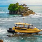 Yellow passenger boat glides near a rocky island with green trees and a thatched pavilion on top, in blue ocean.