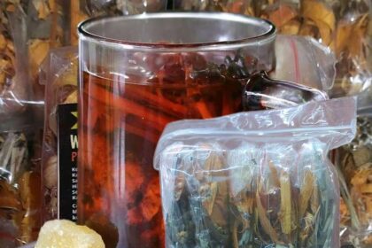 Glass teapot with red herbal tea, alongside a plastic bag of loose tea leaves and a rock sugar on a plate.