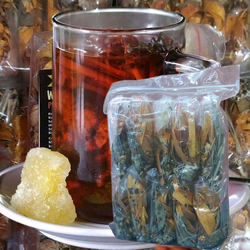Glass teapot with red herbal tea, alongside a plastic bag of loose tea leaves and a rock sugar on a plate.