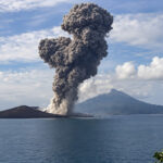 Volcano erupting on a small island, sending a tall ash plume into a blue sky over calm sea.