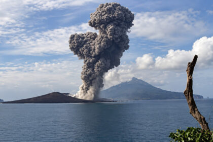 Volcano erupting on a small island, sending a tall ash plume into a blue sky over calm sea.