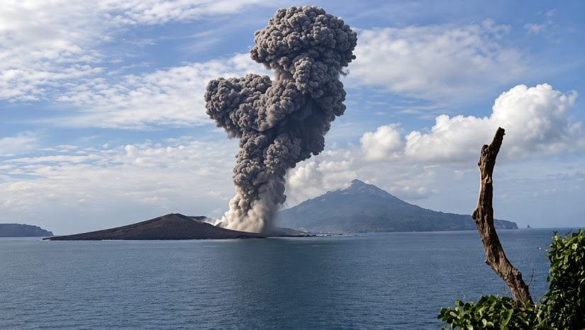 Volcano erupting on a small island, sending a tall ash plume into a blue sky over calm sea.