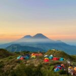Colorful tents scattered on a grassy hillside at sunset with misty mountains in the distance