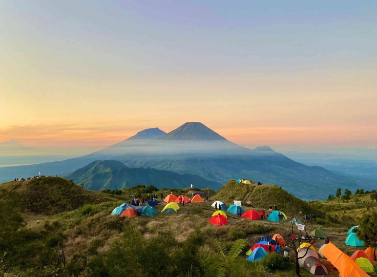 Colorful tents scattered on a grassy hillside at sunset with misty mountains in the distance