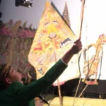 Performer in a green outfit reaches toward colorful, triangular props on stage during a traditional performance.
