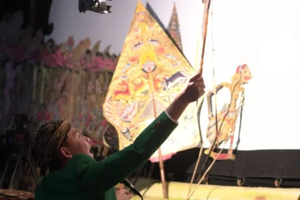 Performer in a green outfit reaches toward colorful, triangular props on stage during a traditional performance.