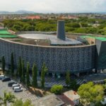 Aerial view of a circular modern building with lattice walls and green rooftop terraces in a tropical city.