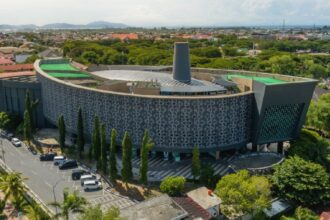 Aerial view of a circular modern building with lattice walls and green rooftop terraces in a tropical city.