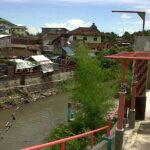 Riverside market area with stalls along a stone embankment, a muddy canal, and a walkway featuring red railings and decorative posts.