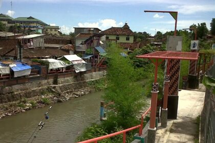 Riverside market area with stalls along a stone embankment, a muddy canal, and a walkway featuring red railings and decorative posts.