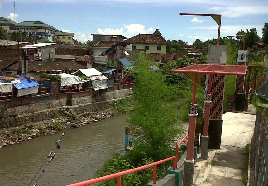 Riverside market area with stalls along a stone embankment, a muddy canal, and a walkway featuring red railings and decorative posts.